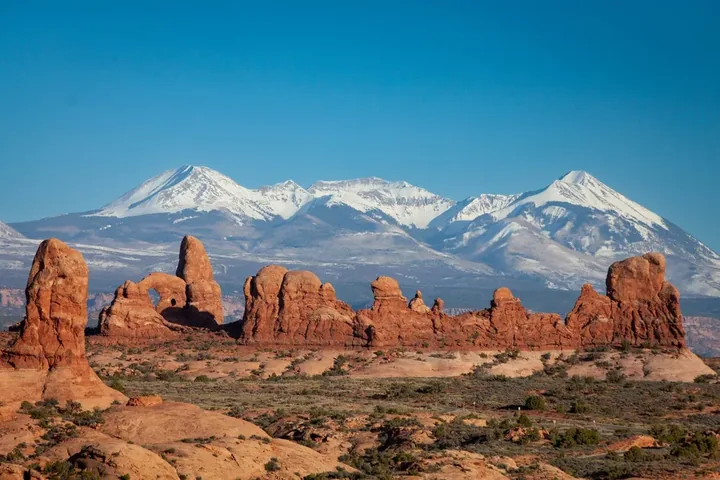 A landscape photo featuring the snow-capped La Sal Mountains in the background, framed by the iconic red rock formations of Arches National Park in the foreground.