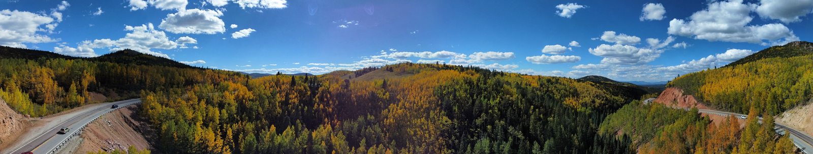 Golden aspens and evergreen hills beside a winding mountain road under a blue, cloud-filled sky.
